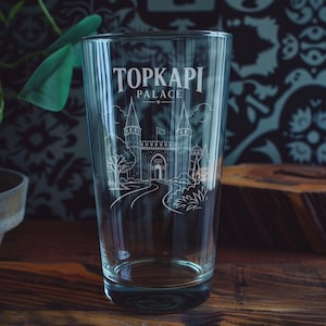 May include: A clear glass pint tumbler featuring a white etched illustration of the Topkapi Palace. The design includes the text TOPKAPI PALACE above a detailed line drawing of the palace gates, towers, and surrounding landscape elements.