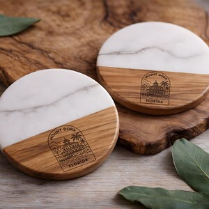 May include: Two round coasters with a white marble top and a wooden base. The wooden base has a Mount Dora, Florida, design. The coasters are on a wooden surface with green leaves.