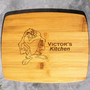 May include: A brown bamboo cutting board with a black border. The board is engraved with a cartoon image of Taz the Tasmanian Devil and the text "VICTOR'S Kitchen".