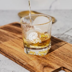 May include: A clear glass with a snake design and the text "Your Custom Text Here" is filled with amber liquid and a large ice cube. The glass is sitting on a wooden cutting board.