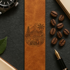 May include: A brown leather bookmark with a detailed engraved design featuring a temple, palm trees, and a jeep. The bookmark is rectangular and lies on a dark surface, surrounded by coffee beans, a watch, and a pen.