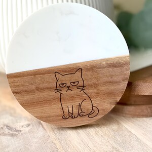 May include: A round coaster with a two-tone design. The top half is white, and the bottom half is wood-toned. A black illustration of a grumpy cat is etched into the wood. The coaster is likely made of wood and resin.