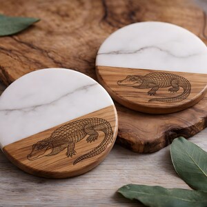 May include: Two round coasters with a half-and-half design. The top half is white marble, and the bottom half is wood with a detailed alligator engraving. The coasters are on a wooden surface, with green leaves in the background.