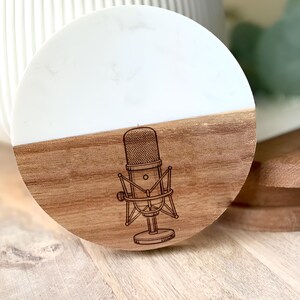 May include: Round coaster with a two-tone design. The top half is white, and the bottom half is wood-toned. A detailed illustration of a vintage microphone is etched into the wood. The coaster is designed for use with beverages.