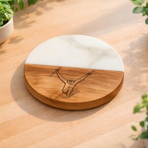 May include: A circular coaster featuring a split design with one half made of white marble and the other half made of natural brown wood. The wooden section is engraved with a detailed line art illustration of a monkey hanging from a branch.