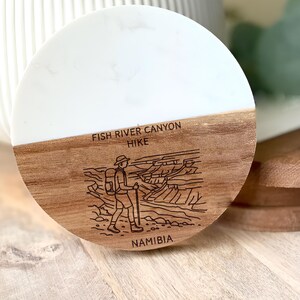 May include: Round coaster with a two-tone design. The top half is white, and the bottom half is wood-toned. The coaster features an etched illustration of a hiker and the text "FISH RIVER CANYON HIKE NAMIBIA."