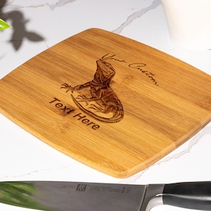 May include: A light brown square-shaped bamboo cutting board with a laser-engraved image of a lizard and the text "Your Custom Text Here".