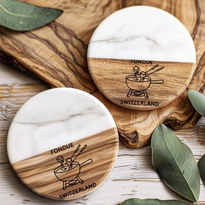 May include: Two round coasters with a white marble top and a wooden bottom. The wooden section has the word "FONDUE" and an illustration of a fondue pot with the word "SWITZERLAND" below it. The coasters are on a wooden cutting board.