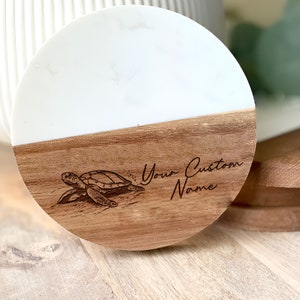May include: A round wooden coaster with a white marble top. The coaster has a laser-engraved image of a sea turtle and the text "Your Custom Name".