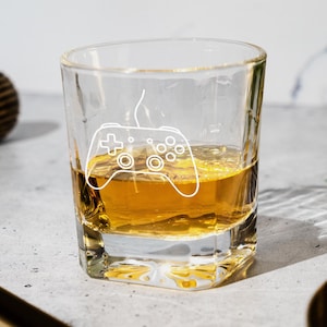May include: A clear, square-shaped glass with a white etched design of a video game controller. The glass is filled with a golden-colored liquid, likely a spirit. The glass sits on a light-colored surface, with a decorative gold ball in the background.