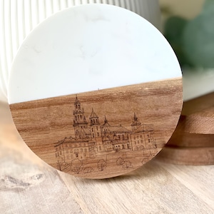 May include: A round coaster with a white top and a wood-grain bottom. The bottom features an etched illustration of a castle. The coaster is a decorative item for home decor.