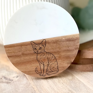 May include: A round wooden coaster with a white top half and a brown bottom half. A detailed line drawing of a cat is etched into the wood. The coaster is sitting on a wooden surface.