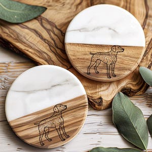 May include: Two round coasters. Each coaster has a white marble top half and a wood-grain bottom half. A black line drawing of a dog is on the wood portion of each coaster. The coasters are on a wooden surface.