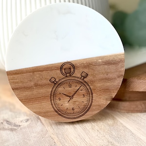 May include: A round coaster with a stopwatch design. The top half is white, and the bottom half is wood. The stopwatch is etched into the wood. The coaster is made of wood and resin.