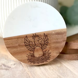 May include: A round coaster with a white top and a wooden bottom. The wooden portion features a detailed engraving of a phoenix in a nest. The coaster is designed to protect surfaces from drink condensation and heat.