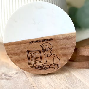 May include: A round coaster with a white top and a wooden bottom. The bottom half features a cartoon illustration of a software engineer working on a computer, with the text "SOFTWARE ENGINEER" above the illustration.