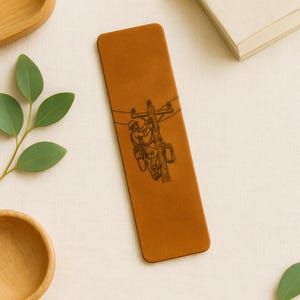 May include: A brown leather bookmark featuring a detailed engraving of a lineman working on a utility pole. The bookmark is rectangular with rounded corners, and the image is etched in a dark brown color.