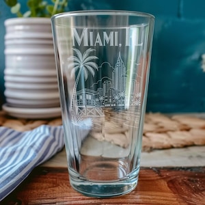 Puede incluir: Un vaso de vidrio transparente con un diseño grabado en blanco de líneas que muestra el horizonte de Miami, FL, incluyendo una palmera y una torre de salvavidas. El texto MIAMI, FL está grabado en una fuente sans-serif en la parte superior.