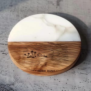 May include: A round coaster with a half-and-half design. The top half is white with a marbled effect, and the bottom half is wood-grained with an etched desert scene. The text "CHEBBI DUNE" is etched along the bottom edge.