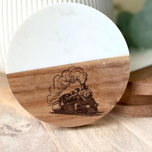 May include: A round coaster with a two-tone design. The top half is white, and the bottom half is wood-toned. A detailed engraving of a vintage steam train is on the wood portion. The coaster is likely used for drinks.