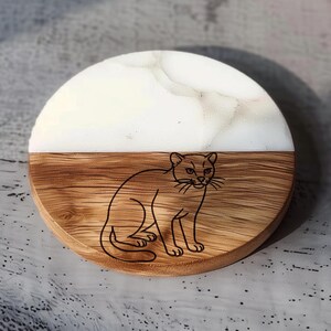 May include: Round wooden coaster with a cat illustration. The top half is white with a marble effect, and the bottom half is wood-grained. A black outline of a cat is etched into the wood.