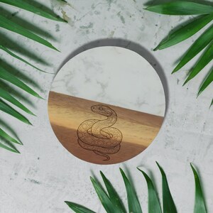 a round, white plate with a black snake design on it, surrounded by green leaves.