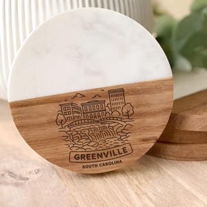 May include: A round coaster with a white marble top and a wooden base. The wooden portion features an engraved illustration of a cityscape with the text "GREENVILLE SOUTH CAROLINA".