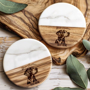 May include: Two round coasters with a white marble top and a wooden bottom. Each coaster features a dog portrait and the text "Your Custom" and "TEXT HERE". The coasters are on a wooden surface.