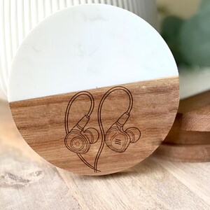 May include: A round coaster with a two-tone design. The top half is white, and the bottom half is wood-toned. A detailed line drawing of in-ear headphones is etched into the wood. The coaster is designed for use with beverages.