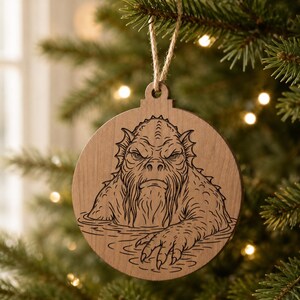May include: Wooden Christmas ornament featuring a creature design. The ornament is round with a cutout at the top for hanging. The creature is depicted in a detailed, engraved style, with a stern expression and clawed hand. The ornament hangs from a natural twine string.