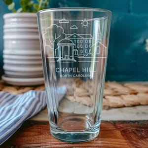 May include: A clear pint glass with a white etched design of a building, tree, and sun, with the words "Chapel Hill North Carolina". The glass is on a wooden surface with a blue and white striped cloth.