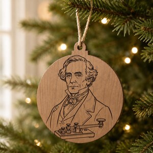 May include: A round wooden Christmas ornament featuring a portrait of Samuel Morse and a telegraph machine. The ornament is suspended by a natural twine string and hangs against a backdrop of a Christmas tree.