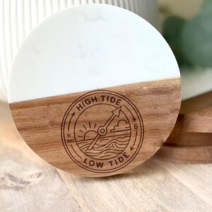 May include: A round coaster with a white top and a wooden bottom. The wooden portion features a circular design with the words "HIGH TIDE" and "LOW TIDE" around a tide clock graphic. The coaster is a decorative item.