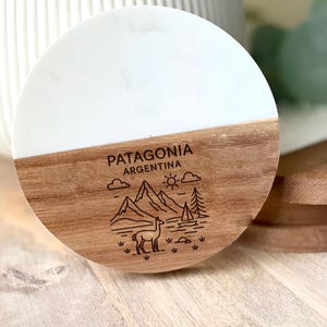 May include: A round coaster with a white top and a wooden bottom. The wooden section is engraved with the text "PATAGONIA ARGENTINA" and a mountain and llama illustration. The coaster is on a wooden surface.
