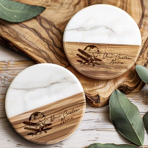 May include: Two round wooden coasters with a white marble top. The bottom half of each coaster is made of wood and features a laser-engraved image of a vintage airplane with the text "Your Custom Name".