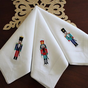 May include: Three white linen napkins with embroidered nutcracker designs. Each napkin features a different nutcracker in a red, blue, and green color scheme.