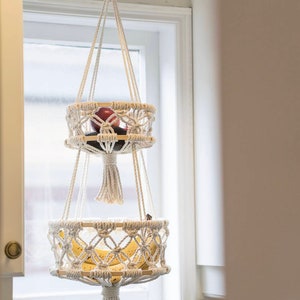 May include: A white macrame hanging fruit basket with two tiers. The top tier holds a red apple and a dark green avocado. The bottom tier holds two yellow bananas. The basket is hanging from a white ceiling hook.