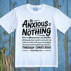 Könnte beinhalten: Weißes T-Shirt mit dem Text "BE ANXIOUS FOR NOTHING" in Schwarz, zusammen mit einem Bibelvers. Das Shirt wird auf einer blauen Holzoberfläche mit getrockneten Pflanzen in der linken unteren Ecke präsentiert.