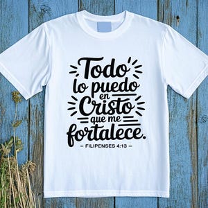 May include: A white t-shirt with the Spanish text "Todo lo puedo en Cristo que me fortalece - Filipenses 4:13" in black script. The shirt is displayed on a blue wooden surface with dried plants.