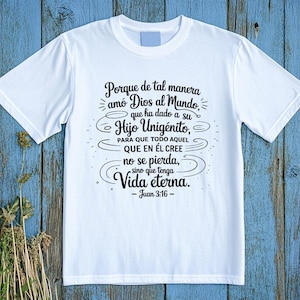 May include: White t-shirt with black text in Spanish. The text includes a religious quote from John 3:16. The shirt is displayed on a blue wooden surface with dried plants in the lower left corner.