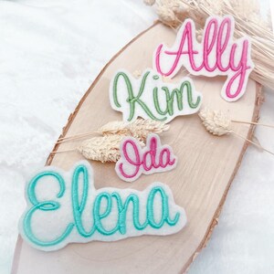 Könnte beinhalten: Vier gestickte Namensaufnäher: "Ally" in Pink, "Kim" in Grün, "Ida" in Pink und "Elena" in Türkis. Die Namen sind auf weißem Filz gestickt und auf einer Holzscheibe mit getrockneten floralen Akzenten angeordnet.