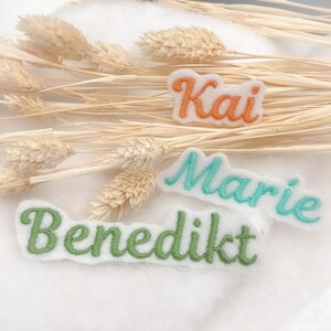Könnte beinhalten: Drei gestickte Namensaufnäher: "Kai" in Orange, "Marie" in Türkis und "Benedikt" in Grün. Die Aufnäher befinden sich auf weißem Hintergrund mit getrockneten Weizenähren.