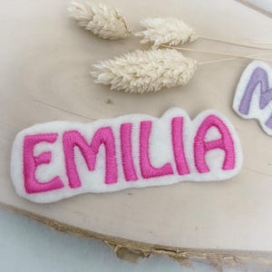 Könnte beinhalten: Ein weißes Filz-Patch mit dem Namen "EMILIA" in leuchtend pinkfarbenen Buchstaben gestickt. Der Aufnäher hat eine abgerundete rechteckige Form. Getrocknete, hellfarbene Blumen sind im Hintergrund.