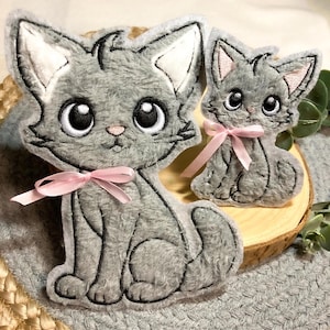 Könnte beinhalten: Zwei graue, katzenförmige Plüschornamente mit schwarzen Umrissen und rosa Schleifen. Die Katzen haben große, ausdrucksstarke Augen und weiße Innenohren. Eines ist größer als das andere. Die Ornamente befinden sich auf einer Holzscheibe und einer grau strukturierten Oberfläche.