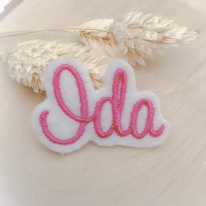 Könnte beinhalten: Ein weißes Filz-Patch mit dem Namen "Ida" in leuchtend pinkfarbener Schreibschrift gestickt. Der Aufnäher hat eine abgerundete rechteckige Form und ist vor einem neutralen Hintergrund mit getrockneten floralen Akzenten platziert.