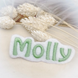 Könnte beinhalten: Ein weißes Filz-Patch mit dem Namen "Molly", der mit grüner Stickerei versehen ist. Der Patch hat eine wolkenartige Form und liegt vor einem weichen, neutralen Hintergrund mit getrockneten Blumen.
