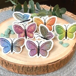 Könnte beinhalten: Eine Sammlung von bunten Schmetterlingsaufnähern mit glitzernden Flügeln. Die Aufnäher sind in verschiedenen Farben wie Blau, Rosa, Orange, Grün und Lila gehalten und auf einer Holzscheibe angeordnet. Jeder Schmetterling hat eine schwarze Umrandung und einen weißen Rand.
