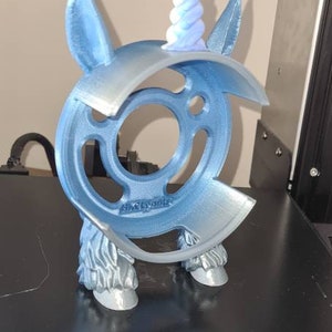 Peut inclure: Une figurine d'une licorne bleue imprimée en 3D avec une corne en spirale bleu clair et violette. La figurine est posée sur une plateforme noire avec le mot "CREALITY" imprimé dessus.