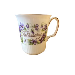 May include: A white porcelain mug with a ribbed base and a gold-trimmed handle. The front features a floral wreath of purple violets surrounding the word February written in a gold cursive script. The rim of the mug is also finished with gold.