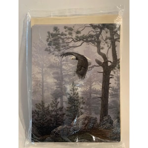 May include: A stack of rectangular greeting cards enclosed in a clear plastic sleeve. The top card features a detailed illustration of a bald eagle in flight over a misty forest landscape with tall pine trees and large grey rocks in the foreground.
